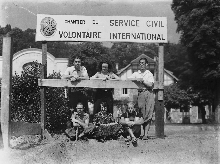 Vieille photo en noir et blanc. Des volontaires adultes se tiennent debout devant une palissade surmontée du logo du service civil international. Deux hommes et une femme au centre sont assis par terre. Deux hommes et une femme au centre se tiennent debout derrière eux.
Le paysage derrière eux présente de multiples sapins et ce qui semble être des maisons.