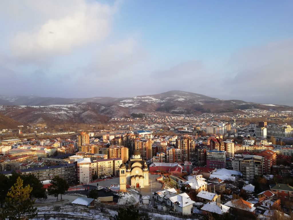 Vue panoramique de la ville de Mitrovica au couché du soleil. Au centre on observe une église, autour des tours d'immeubles résidentielles. Au fond, un paysage montagneux.